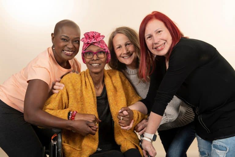 Four women embrace and smile at the camera. One woman is presumably in treatment for breast cancer and sits in a wheelchair with a pink scarf on her head.