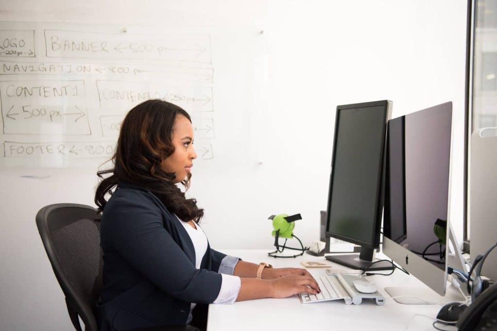 A woman sits at a desktop computer, typing. On the whiteboard behind her is a wireframe for a webpage.