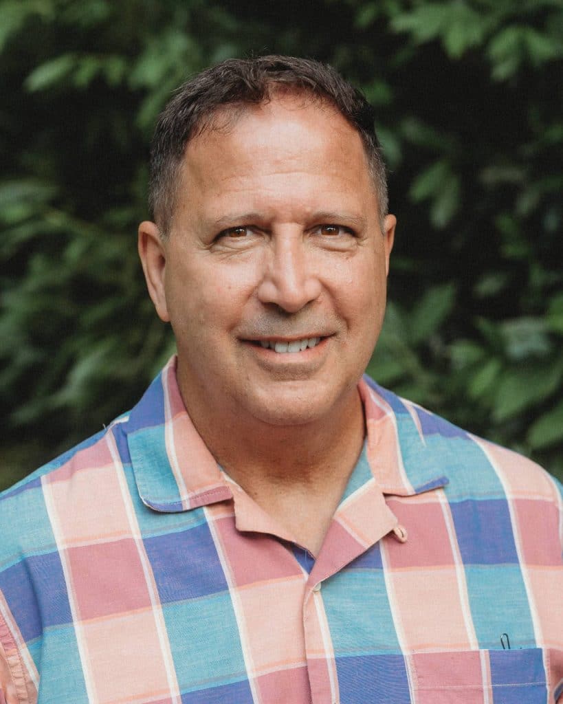 An outdoor headshot of Bob Scavilla, the CEO of FourFront, wearing a blue and pink checkered shirt in front of a leafy green background.