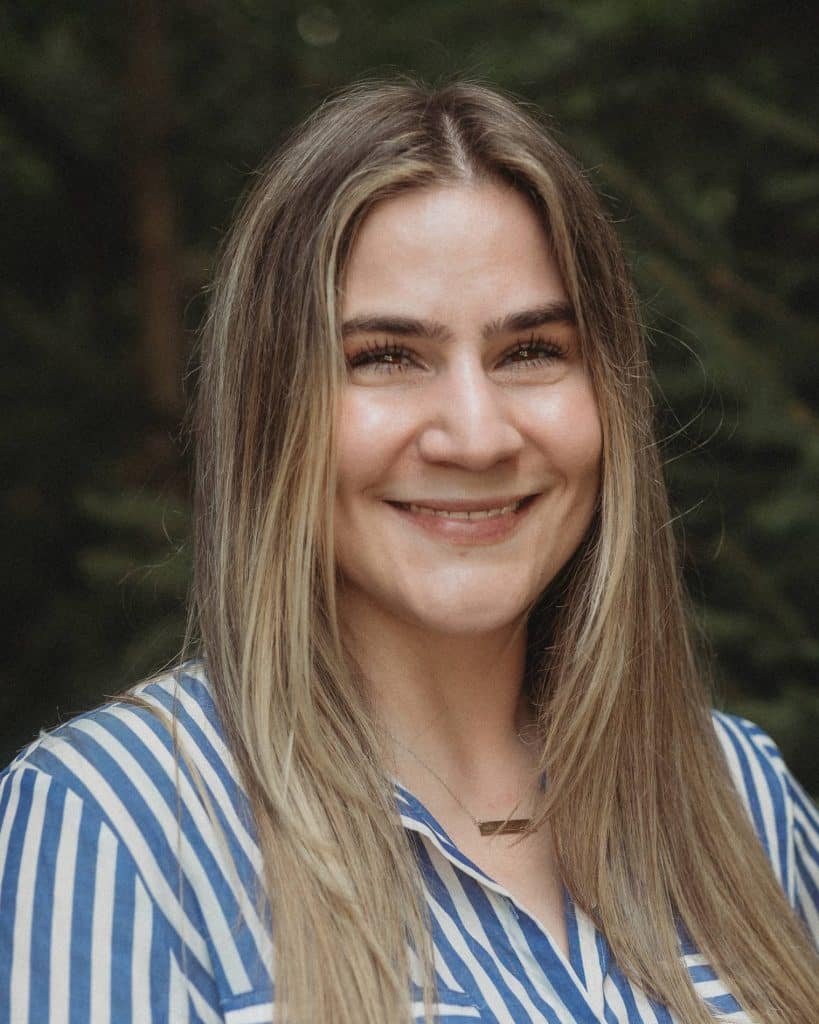 An outdoor headshot of Christina Lewis wearing a striped shirt and smiling in front of a leafy green background.