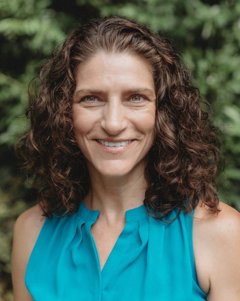 An outdoor headshot of Kim Kalishek, the CFO of FourFront, wearing a bright blue shirt and smiling in front of a leafy green background.