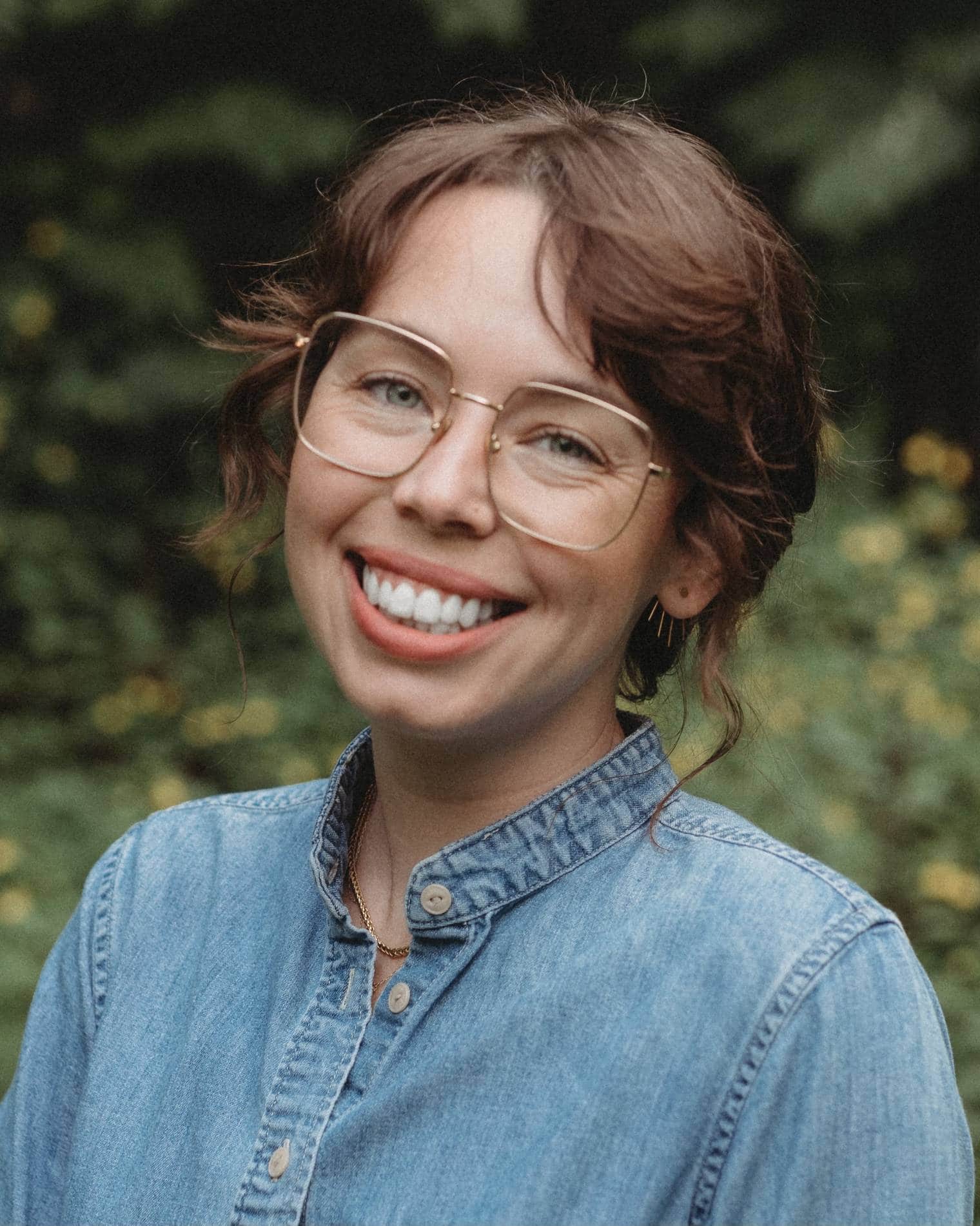 Leah MacDaniel, smiling and wearing a denim shirt and gold glasses.